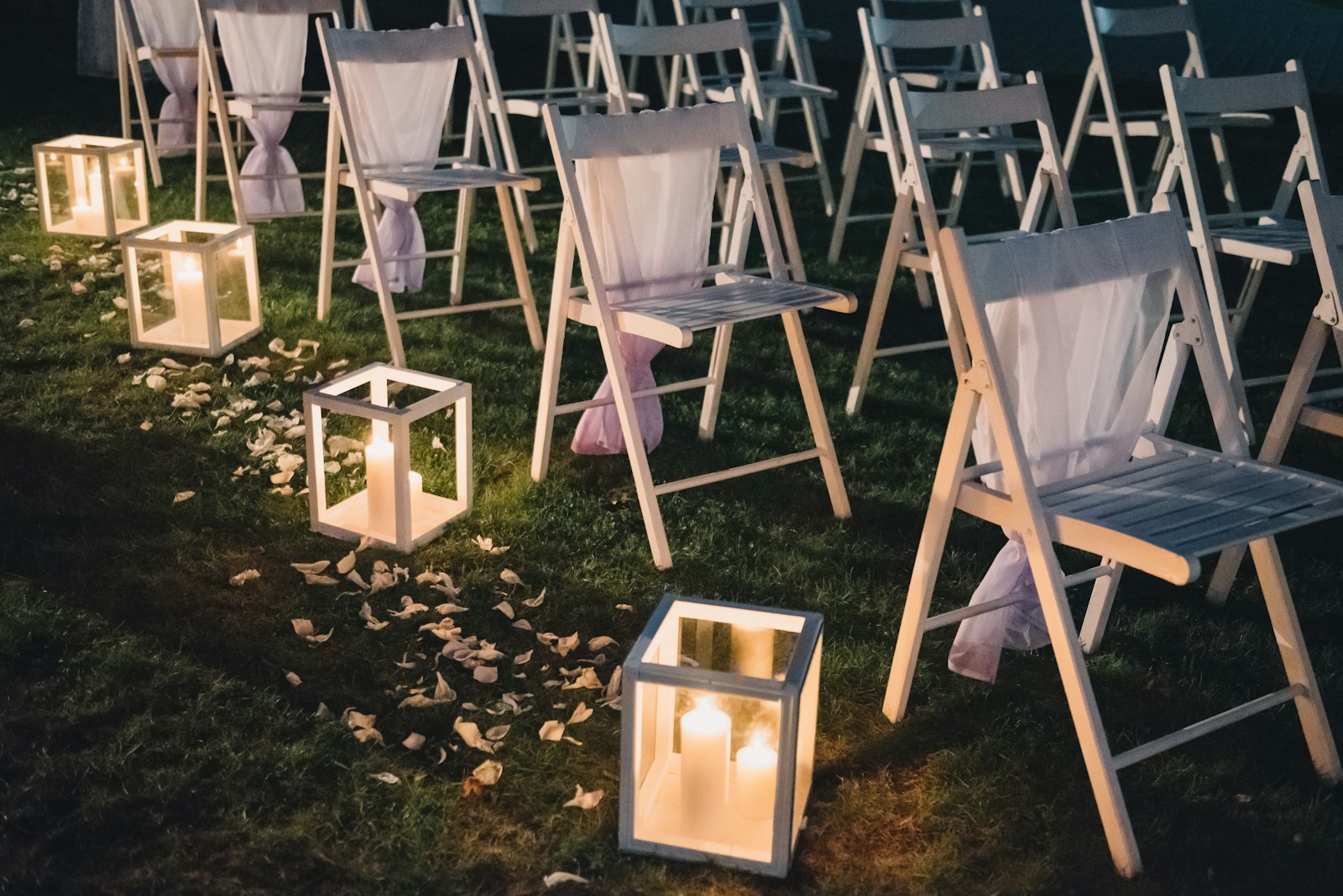Candle lanterns and scattered petals along an aisle of white folding chairs at dusk