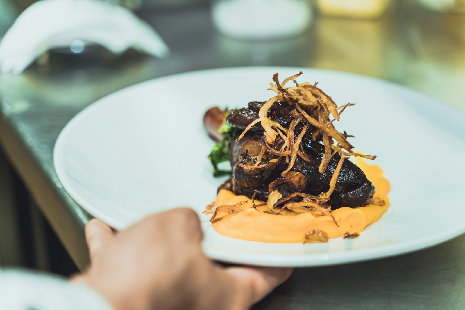 Braised beef topped with crispy shallots on golden polenta, the plate held at a restaurant kitchen pass