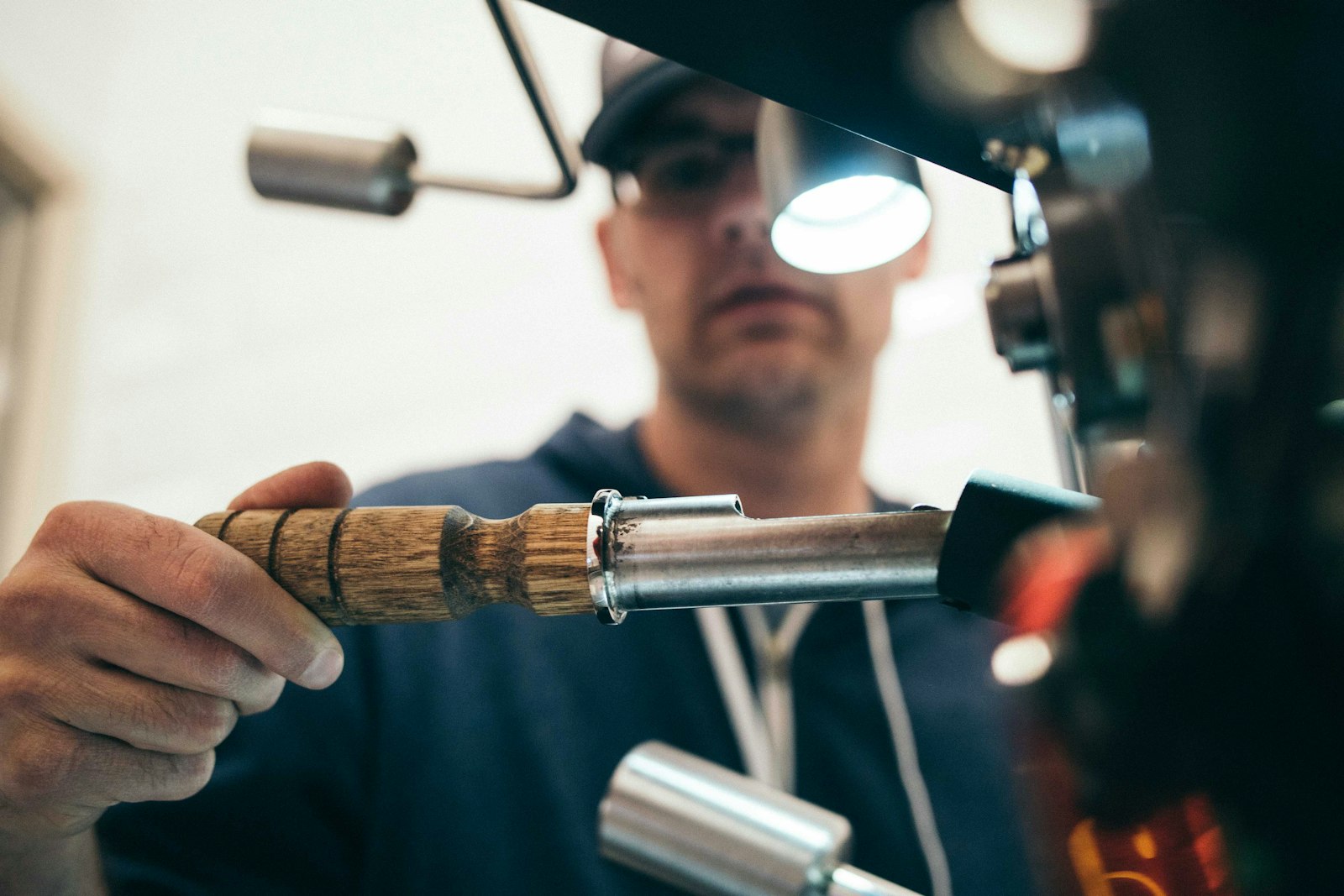 Man in a cap and glasses pulling a wooden-handled sampler from a coffee roaster