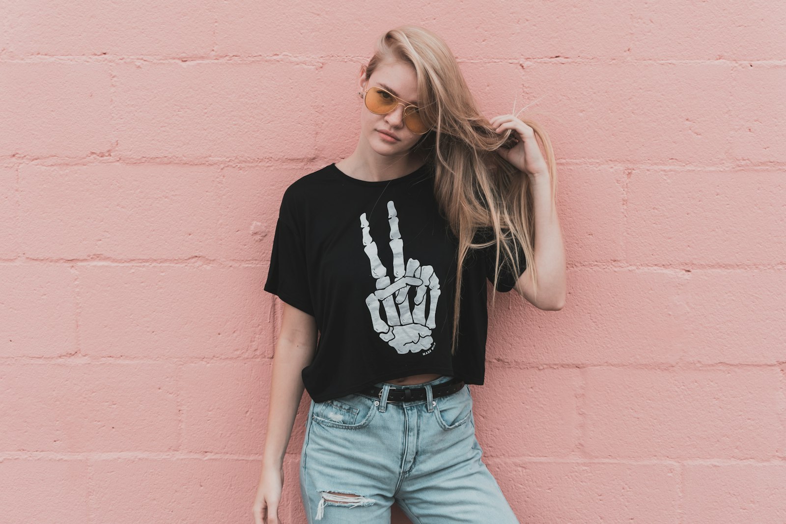 Blonde woman in sunglasses and a skeleton-hand tee against a pink brick wall