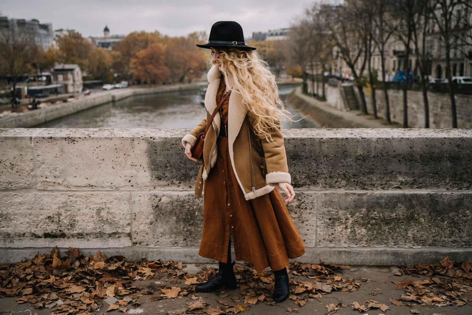 Woman in a long tan suede coat and black hat on a stone bridge over a river, autumn leaves at her feet